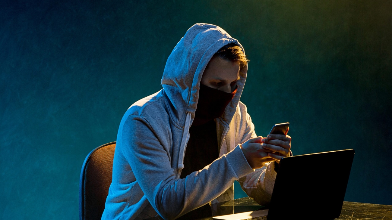 Hooded computer hacker stealing information with laptop on colored studio background